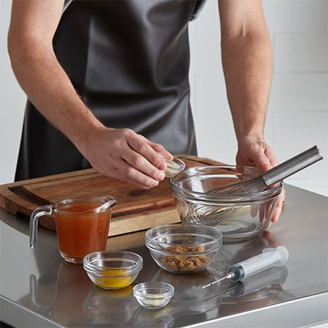 A chef adding onion powder, garlic powder, brown sugar, mustard, and apple cider to a mixing bowl.