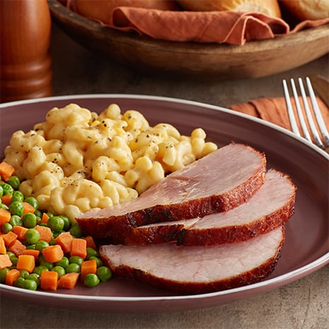 A dinner plate with three slices of ham, mac and cheese, peas and carrots