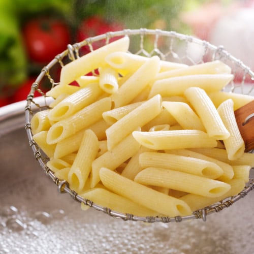 A scoop of penne pasta being drained over a boiling pot