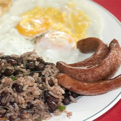 a white plate with two fried eggs, strips of sliced sausage, and a serving of rice and beans
