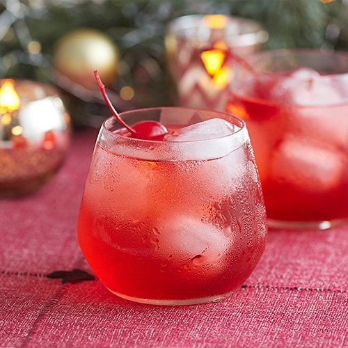 A glass of a drink that is red, with ice cubes and a cherry called a Red Stag Dirty Shirley.
