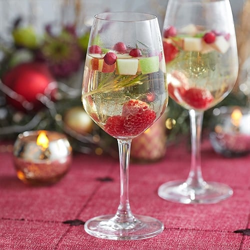 Two elegant glasses of a drink with raspberries, cranberries, cubed apples, and a rosemary sprig.