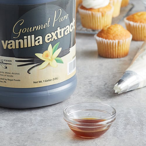 Container of vanilla flavor extract surrounded by small glass bowl of vanilla extract and unfrosted cupcakes