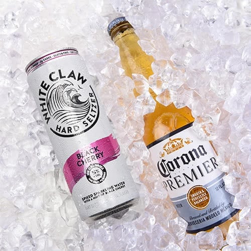can of White Claw black cherry hard seltzer laying in ice beside a bottle of Corona beer