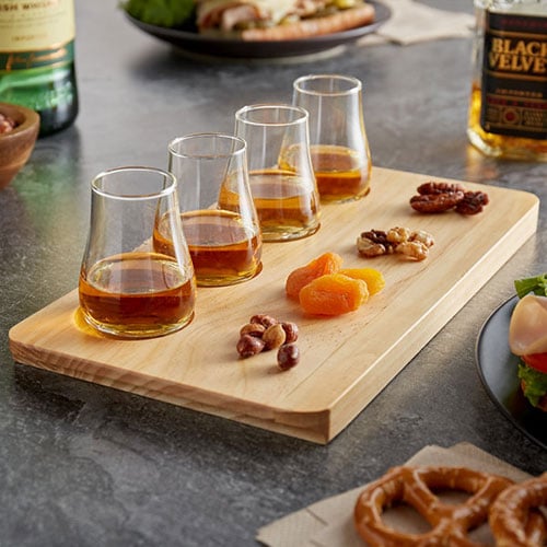 four glasses of whiskey on wooden flight with pecans hazelnuts walnuts dried fruit and pretzels
