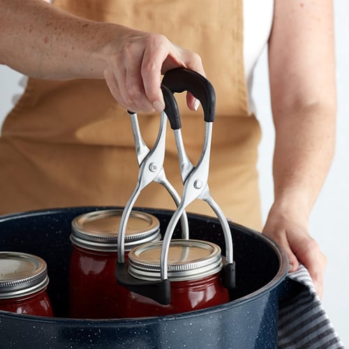 person lifting a hot sauce jar out of pot with jar lifter