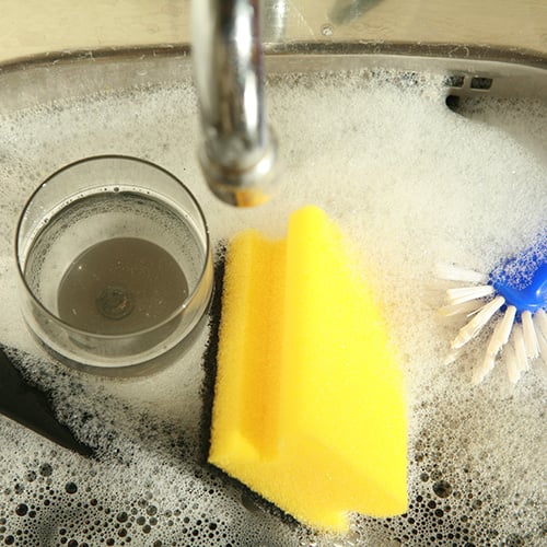a glass and sponge soaking in a sink filled with a soapy water.