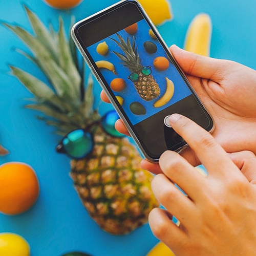 hands holding phone and taking photo of stylish pineapple in sunglasses with fruit in background