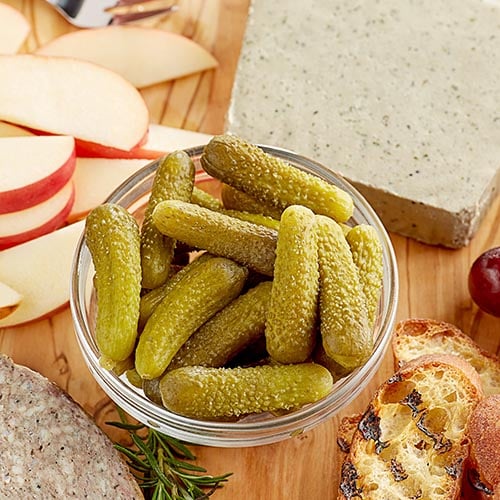 dill pickles in round glass bowl on wooden table with toasted baguette apples and cheese