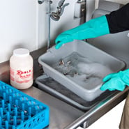 Gloved hands holding a tub of stainless steel flatware soaked in sanitizing solution