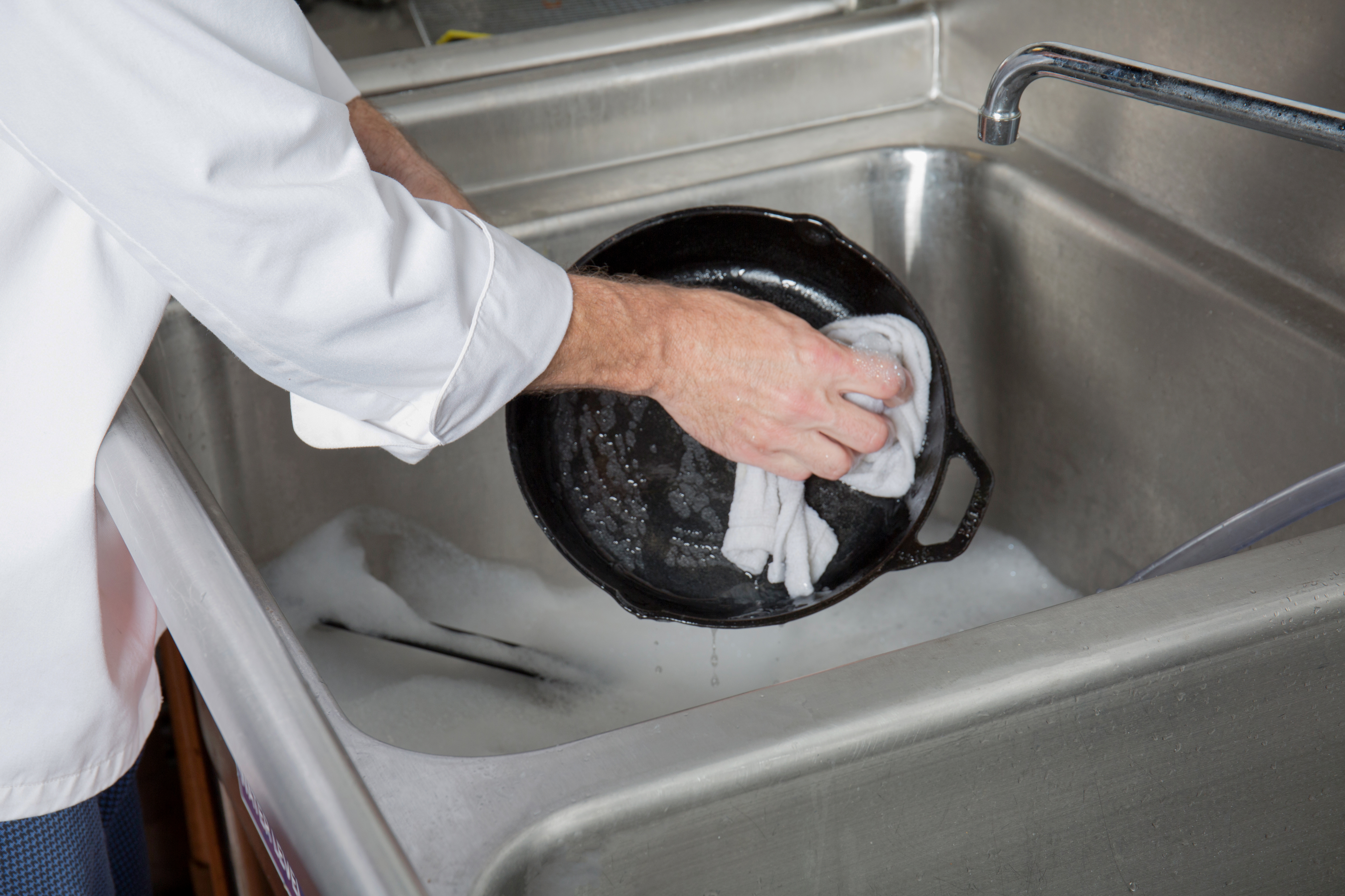 Chef washing a cast iron skillet in sink with soapy water and wash cloth